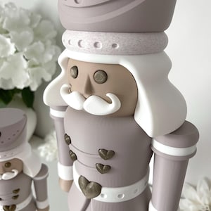 May include: Two nutcracker figures in a muted color palette of taupe, white, and metallic bronze. The figures feature tall hats with heart accents, white hair, white mustaches, and jackets adorned with small bronze heart-shaped buttons.