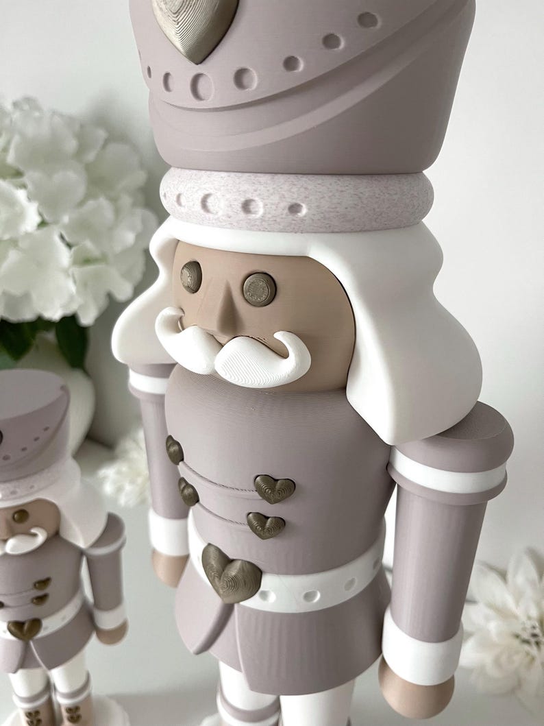 May include: Two nutcracker figures in a muted color palette of taupe, white, and metallic bronze. The figures feature tall hats with heart accents, white hair, white mustaches, and jackets adorned with small bronze heart-shaped buttons.
