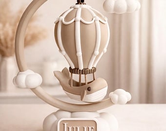 May include: A decorative nursery mobile featuring a hot air balloon design with a whale-shaped basket. The mobile is in neutral tones of beige and white, with the name "JULIE" displayed on a cloud-shaped base.
