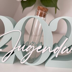 May include: A light blue decorative sign with the number "2022" and the word "Jugend" in white script. A clear glass vial with a cork stopper is placed in the center of the sign. White flowers are in the background.