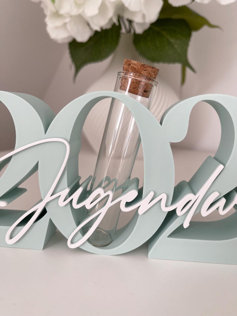 May include: A light blue decorative sign with the number "2022" and the word "Jugend" in white script. A clear glass vial with a cork stopper is placed in the center of the sign. White flowers are in the background.