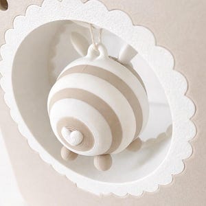 May include: A beige and white striped decorative object, possibly a mobile or ornament, hangs within a scalloped-edged frame. The object has a heart-shaped detail and small feet. The frame is a light beige colour.