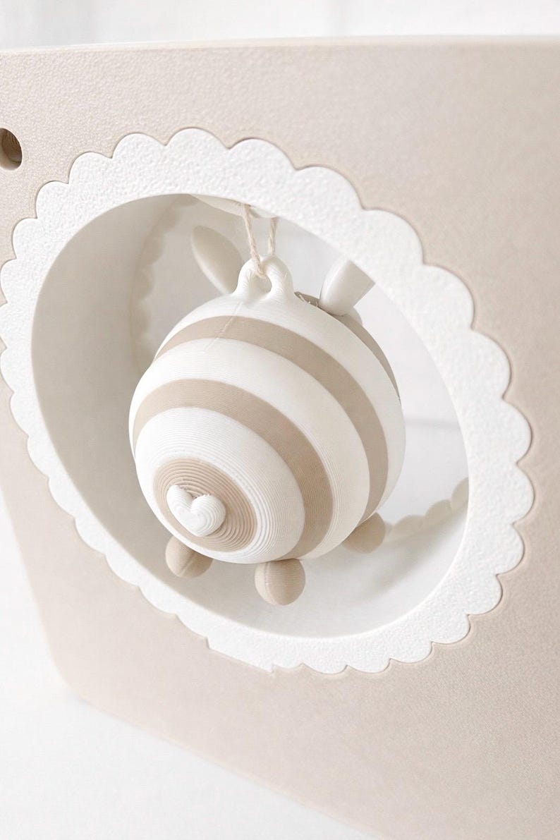 May include: A beige and white striped decorative object, possibly a mobile or ornament, hangs within a scalloped-edged frame. The object has a heart-shaped detail and small feet. The frame is a light beige colour.