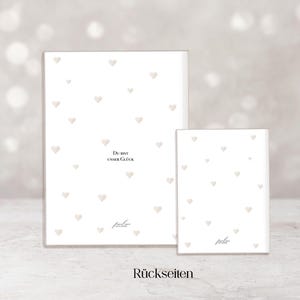 May include: Two white rectangular cards with a pattern of small, light-colored hearts. The larger card has the text "Du bist unser Gl&uuml;ck" and the smaller card has the text "R&uuml;ckseiten". The cards have a soft, neutral background.