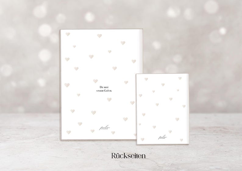 May include: Two white rectangular cards with a pattern of small, light-colored hearts. The larger card has the text "Du bist unser Gl&uuml;ck" and the smaller card has the text "R&uuml;ckseiten". The cards have a soft, neutral background.