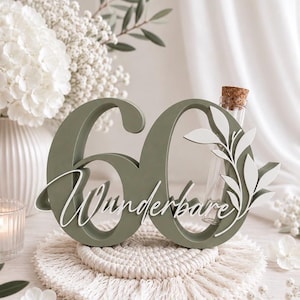 May include: A sage green wooden number 60 sign featuring the white script text Wunderbare and a white leaf cutout design. A clear glass test tube with a cork stopper is integrated into the number zero, providing a space for small floral stems.