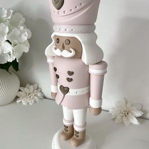 May include: A standing nutcracker figurine featuring a soft pink and white color palette. The figure wears a tall pink hat with a bronze heart emblem, a pink jacket with bronze heart buttons, white trousers, and tan boots with bronze heart accents.