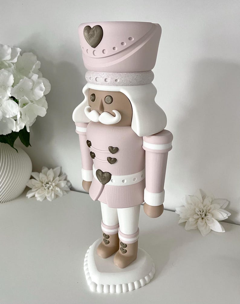 May include: A standing nutcracker figurine featuring a soft pink and white color palette. The figure wears a tall pink hat with a bronze heart emblem, a pink jacket with bronze heart buttons, white trousers, and tan boots with bronze heart accents.
