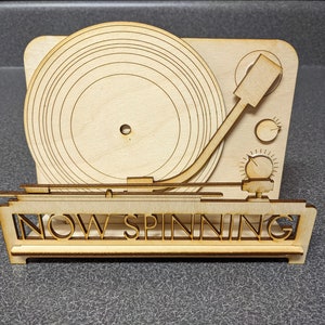 May include: A wooden cutout of a record player with the words "NOW SPINNING" on the front. The record player is made of light wood and has a simple design.