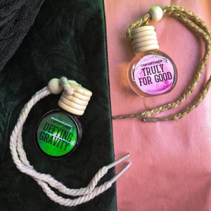 May include: Two car air fresheners with wooden tops and hanging cords. One has a green label with "Defying Gravity" text, the other has a pink label with "Truly For Good" text. The background is a mix of green and pink.