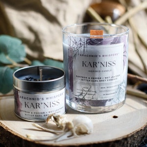 May include: Two candles with a spiderweb design and the text "Arachnid's Whispers Kar'niss Inspired Candle Saffron & Pepper Wet Leather Bitter Earl Grey Essence Vegan 100% Organic Soy Wax Handmade in Germany 300 ml - 50 hrs Burning By Kaleidoneon"