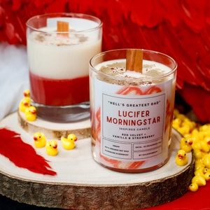 Puede incluir: Dos velas en capas rojas y blancas en frascos de vidrio con una mecha de madera. La vela de la derecha tiene una etiqueta que dice "Hell's Greatest Dad" y "Lucifer Morningstar Inspired Candle Red Velvet Vanilla & Strawberry Vegan 100% Organic Soy Wax Handmade in Germany 300 ml - 50 hrs Burning Time By Kalieignew".
