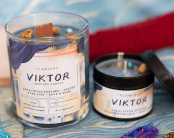 Könnte beinhalten: Zwei Kerzen mit blauen und weißen Etiketten. Die Etiketten lauten "Flawless" Viktor Inspired Candle" und listen die Düfte als "Eukalyptus-Essenz, Goldener Sternanis, Salbei & Moschus" auf. Die Kerzen befinden sich in Glasbehältern und haben ein blaues Wachs mit goldenen und braunen Akzenten.