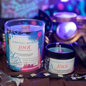 Op de afbeelding: Twee blauwe en roze kaarsen met de tekst "Bombshell Madness Jinx Inspired Candle Popping Bubblegum Cotton Candy" op het etiket. De kaarsen bevinden zich in glazen potten met zwarte deksels.