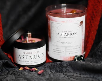 May include: Two candles with black lids and labels that read "Blood Lust Astarion. Inspired Candle" and "Blood Lust Astarion" with the scent notes "White Musk - Black Lily and Clean Amber".