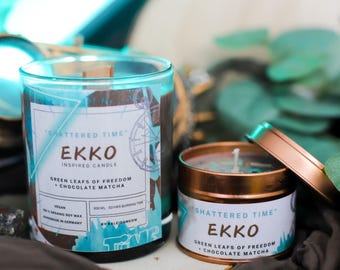 Könnte beinhalten: Zwei Kerzen im Teal- und Kupferdesign. Die Kerzen sind mit "Shattered Time" und "Ekko Inspired Candle" beschriftet. Der Duft ist "Green Leafs of Freedom + Chocolate Matcha".