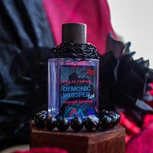 Könnte beinhalten: Ein rechteckiger Parfümflakon mit dunklem Holzdeckel und schwarzer Kette um den Hals. Auf dem Etikett steht "DEMONIC WHISPER" und "INSPIRED PERFUME". Die Flasche steht auf einem Holzblock, umgeben von schwarzen Perlen.