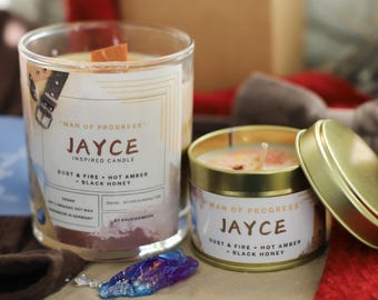 Könnte beinhalten: Zwei goldene Kerzen mit dem Etikett "Man of Progress" und dem Namen "Jayce" darauf. Die Kerzen sind mit "Dust & Fire + Hot Amber + Black Honey" beschriftet. Die größere Kerze befindet sich in einem durchsichtigen Glasgefäß und die kleinere Kerze in einer goldenen Blechdose.