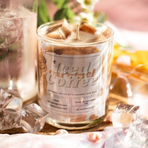 Könnte beinhalten: Ein Glas-Kerze mit der Aufschrift "Iced Caramel Coffee". Die Kerze enthält hellbraunes Wachs und einen Holzdocht. Das Etikett weist darauf hin, dass sie aus Bio-Sojawachs hergestellt wurde und ein Volumen von 300 ml hat. Eiswürfel sind verstreut.