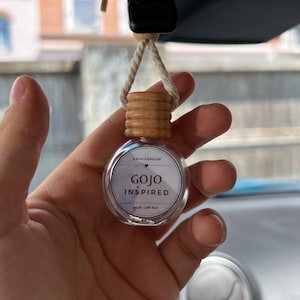 Könnte beinhalten: Eine durchsichtige Glasflasche mit einem Holzdeckel und einem weißen Etikett mit der Aufschrift "Kaleidomeow Gojo Inspired made with love". Die Flasche hängt an einer Schnur und wird von einer Hand gehalten.