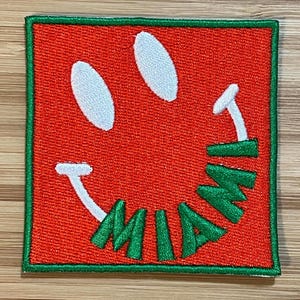 May include: A red and green embroidered patch with a smiley face and the word "MIAMI" written in a curved line around the bottom of the face.