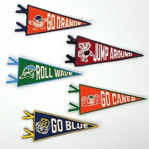May include: Five embroidered pennants in orange, red, green, and blue. Each pennant features team-related text and mascot graphics. The pennants are triangular with decorative edges and are likely intended for display or as patches.