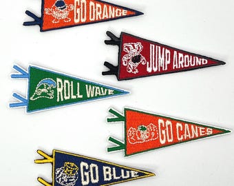 May include: Five embroidered pennants in orange, red, green, and blue. Each pennant features team-related text and mascot graphics. The pennants are triangular with decorative edges and are likely intended for display or as patches.