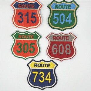 May include: Five embroidered patches in the shape of road signs. Each patch features the word "ROUTE" above a number: 315, 504, 305, 608, and 734. The patches have various color combinations, including red, green, blue, and yellow.
