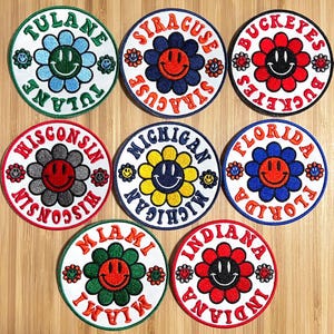 May include: Nine embroidered patches with a smiley face flower design. Each patch features a different university name, including Tulane, Syracuse, Buckeyes, Wisconsin, Michigan, Florida, Miami, and Indiana.