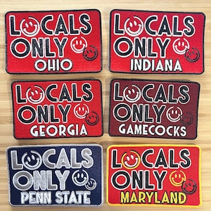 May include: Five red, white, and black embroidered patches with the text "Locals Only" and the state name below. The patches are for Ohio, Indiana, Georgia, Gamecocks, and Maryland.