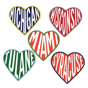 May include: Five heart-shaped embroidered patches. Each patch features a state or university name in white, set against a colorful background. The patches include Michigan, Wisconsin, Miami, Tulane, and Syracuse, with contrasting border colors.
