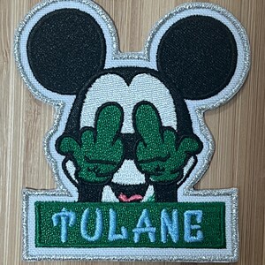 May include: Embroidered patch featuring a cartoon Mickey Mouse with green hands covering his eyes. The patch has a white background with a green banner that reads "TULANE" in blue lettering.