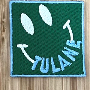 May include: A green embroidered patch with a smiling face design. The face features white oval eyes and a curved white mouth with the word "TULANE" in light blue. The patch has a light blue border.