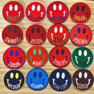 May include: A collection of 16 embroidered smiley face patches with different college names. The patches are red, blue, green, brown, and yellow. The names include: Washington, Wisconsin, Arizona, Trojans, Seminoles, Boston, Texas, Virginia, Florida, Miami, Syracuse, Tulane, Michigan, Villanova, Penn State, and Lehigh.