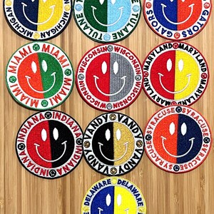 May include: Twelve round embroidered patches with different college names and a smiley face design. Each patch has a different color combination. The patches are on a wooden surface.