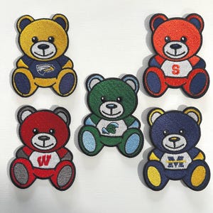 May include: Five embroidered teddy bear patches with different colored fur and team logos. The bears are wearing blue, red, green, yellow, and navy blue shirts with team logos. The logos include a bird, a letter 'S', a wave, a letter 'W', and a letter 'M'.
