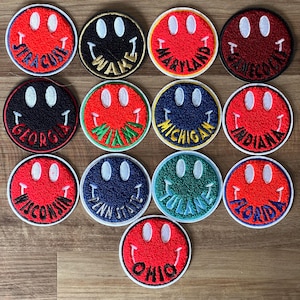 May include: A collection of 14 red, white, and blue smiley face patches with different college names. The patches are made of fuzzy fabric and have a white outline. The names of the colleges are Syracuse, Wake, Maryland,  South Carolina, Georgia, Miami, Michigan, Indiana, Wisconsin, Penn State, Tulane, Florida, and Ohio.