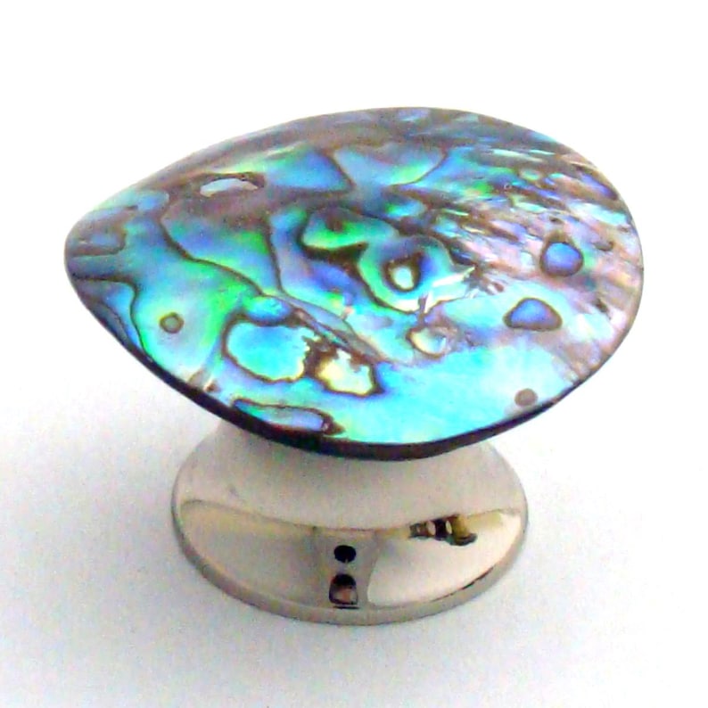 May include: A cabinet knob featuring a polished, iridescent abalone shell inlay with swirling patterns of blue, green, and purple hues. The shell is mounted on a smooth, circular, silver-toned metal base with a reflective finish and a central screw hole.