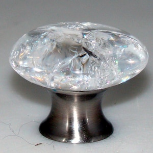 May include: A clear, oval-shaped cabinet knob with a cracked, crystalline appearance. The knob is mounted on a dark, metallic base. The knob's transparent design allows light to pass through, creating a sparkling effect. The base is approximately 1.5 inches in diameter.