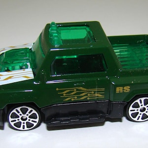 May include: A green and black toy pickup truck with silver and white wheels. The truck has a clear green roof and a white hood with gold circuit board designs. The side of the truck has gold circuit board designs and the letters "RS".