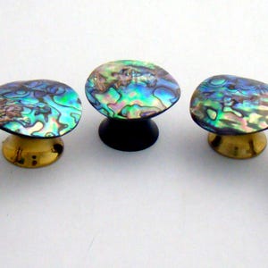 May include: Five circular cabinet knobs featuring iridescent abalone shell tops with swirling patterns of blue, green, and purple. The knobs have varied bases in metallic gold, black, and silver tones, each with a distinct mounting hardware design.