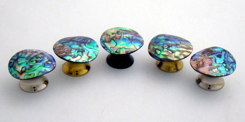 May include: Five circular cabinet knobs featuring iridescent abalone shell tops with swirling patterns of blue, green, and purple. The knobs have varied bases in metallic gold, black, and silver tones, each with a distinct mounting hardware design.