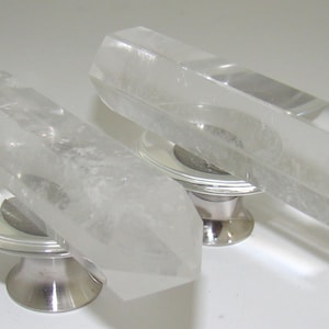 May include: Two cabinet knobs featuring clear, faceted, hexagonal crystal points attached to circular, polished silver-tone metal bases. The crystal elements are elongated and transparent, showcasing natural internal inclusions and sharp, geometric edges.
