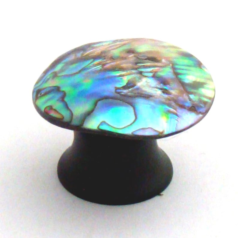 May include: A cabinet knob featuring a round, iridescent abalone shell inlay with swirling patterns of blue, green, and purple hues. The base of the knob is a solid, matte black cylindrical shape that tapers slightly towards the centre.