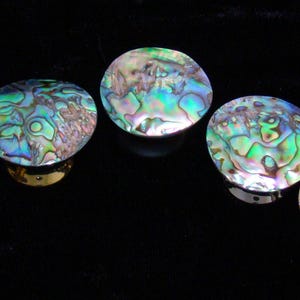 May include: Five circular cabinet knobs featuring iridescent abalone shell inlays with swirling patterns of green, blue, and pink. Each knob has a polished metallic base that supports the rounded, shimmering shell top, creating a reflective surface.