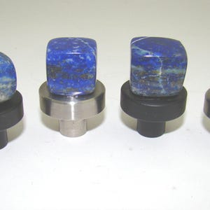 May include: Four cabinet knobs featuring polished, deep blue lapis lazuli stone tops with natural white and gold-toned inclusions. Each stone is mounted on a circular base, with two knobs featuring matte black bases and two featuring brushed silver metal bases.