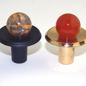 May include: Four cabinet knobs featuring spherical stone tops. Two knobs have matte black bases with green moss agate and brown patterned stone spheres. Two knobs have polished gold bases with a solid red sphere and a dark grey stone sphere.