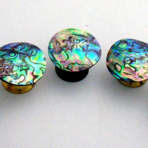 May include: Five circular knobs featuring iridescent abalone shell inlays with swirling patterns of green, blue, and purple. Each knob has a metallic base, with three silver-toned bases and two gold-toned bases visible in this arrangement.