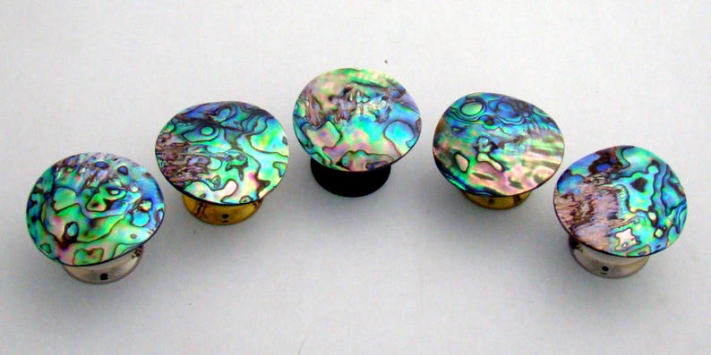 May include: Five circular knobs featuring iridescent abalone shell inlays with swirling patterns of green, blue, and purple. Each knob has a metallic base, with three silver-toned bases and two gold-toned bases visible in this arrangement.