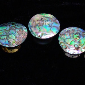 May include: Five circular cabinet knobs featuring iridescent abalone shell inlays with swirling patterns of green, blue, and purple. Each knob has a polished metallic base that supports the rounded, shimmering shell top, creating a reflective surface.